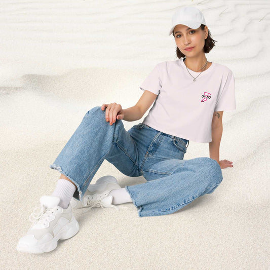 Frau sitzt auf dem Sand und trägt eine blaue Jeans, weiße Sneaker und eine weiße Cap, passend zu dem OH NO crop top t-shirt von wildtides