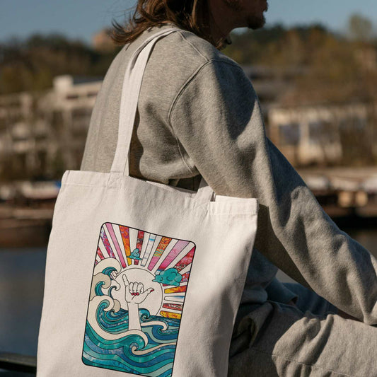 Mann trägt die Shaka Vibes Jute-Tasche von Wild Tides mit buntem Surf-Print im Sonnenuntergang