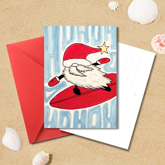Handgezeichnete Weihnachtskarte „Ho Ho Surf“ mit surfendem Weihnachtsmann und rotem Umschlag auf Sandhintergrund