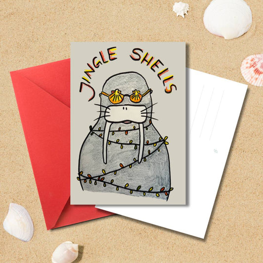 Handgezeichnete Weihnachtskarte „Jingle Shells“ mit Robbe, Lichterkette und rotem Umschlag auf Sandhintergrund